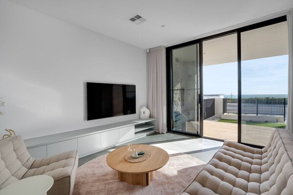 Un salon moderne avec un téléviseur à écran plat fixé au mur, une table basse ronde en bois et une grande porte-fenêtre coulissante donnant sur une terrasse extérieure.