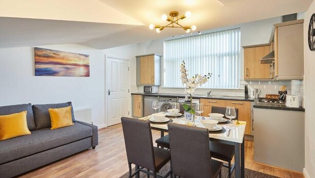 A modern kitchen with white cabinets, a dining table set for four, and a gray sofa with yellow cushions.