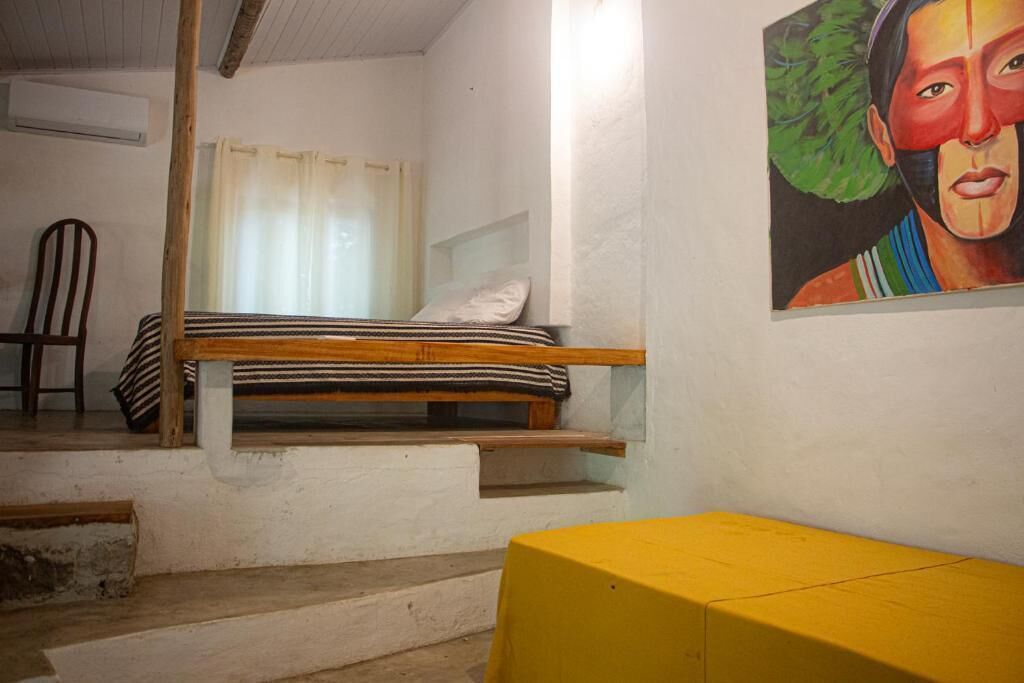 Una stanza con un letto, una sedia, un tavolo giallo e un quadro appeso al muro.