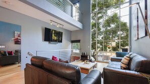 A modern living room with leather sofas, a flat-screen TV, and large windows offering a view of trees.