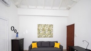 Een kamer met een grijze bank, twee gele kussens, een klein zwart bureau en een abstract schilderij aan de muur.