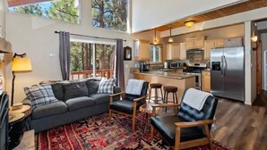 Een ruime woonkamer met een bank, stoelen en een kamerbreed tapijt. Een keuken met moderne apparatuur en een eethoek met krukken is zichtbaar.