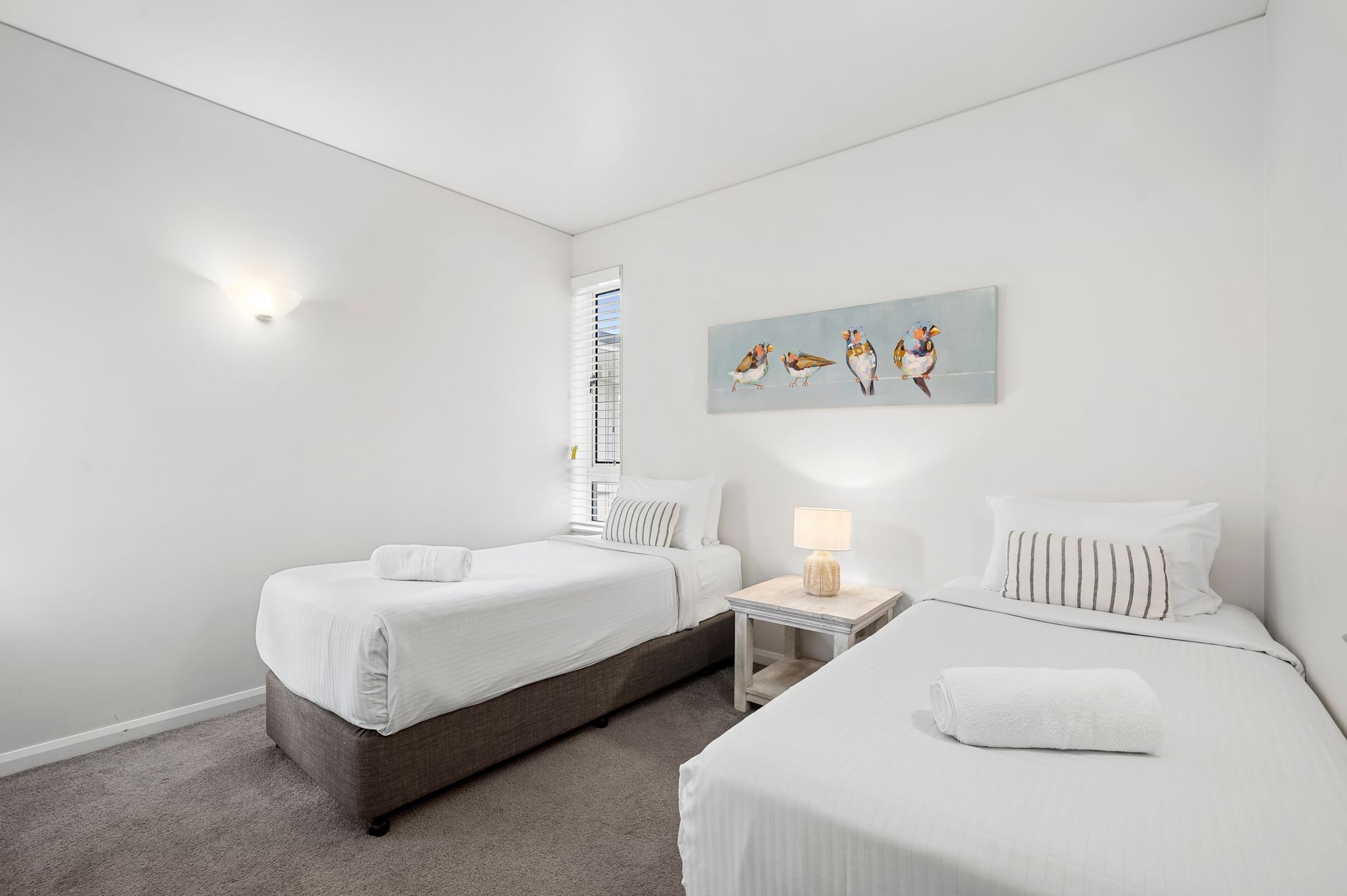 A hotel room with two single beds, a small bedside table with a lamp, a window with blinds, and a wall-mounted artwork of birds.