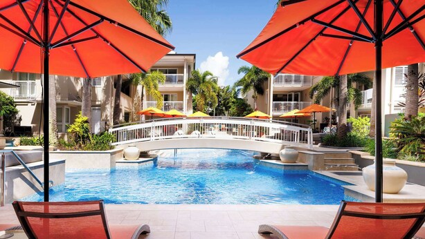 A pool area with lounge chairs and umbrellas, a bridge, and a building with balconies. - Hastings Suites by Stay Noosa (Noosa Heads)