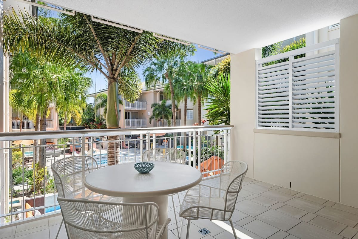 A balcony with a round table and chairs, overlooking a pool and palm trees.