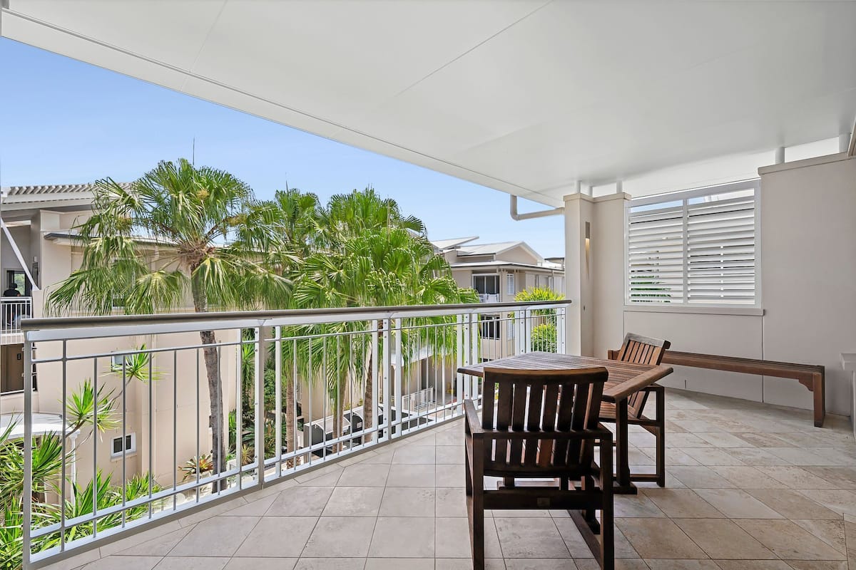 A balcony with a table and chairs, a white railing, and a view of palm trees and buildings.