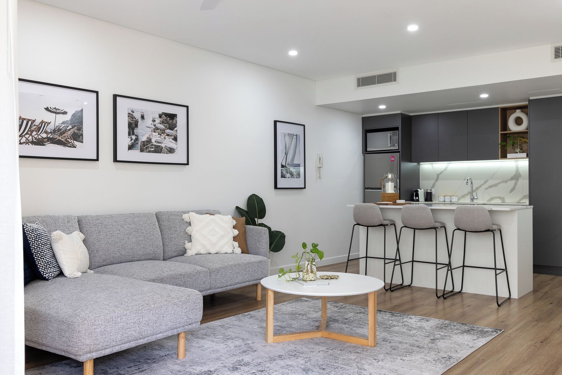 A modern living room with a grey sofa, a white coffee table, and a kitchen area with a bar and stools.