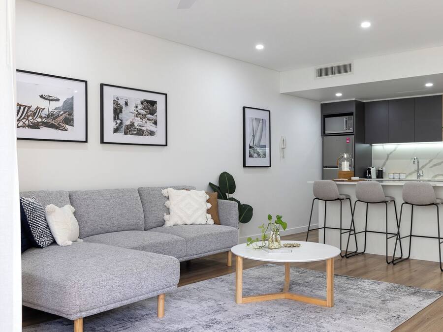 A modern living room with a grey sofa, a white coffee table, and a kitchen area with a bar and stools.