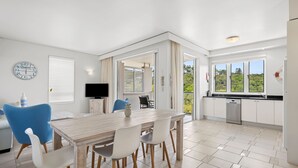 A modern kitchen with a dining area, white cabinetry, and a large window with a view of greenery. - Hastings Suites by Stay Noosa (Noosa Heads)