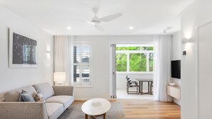A modern living room with a sofa, a small round table, a ceiling fan, and a TV on a stand. - Hastings Suites by Stay Noosa (Noosa Heads)