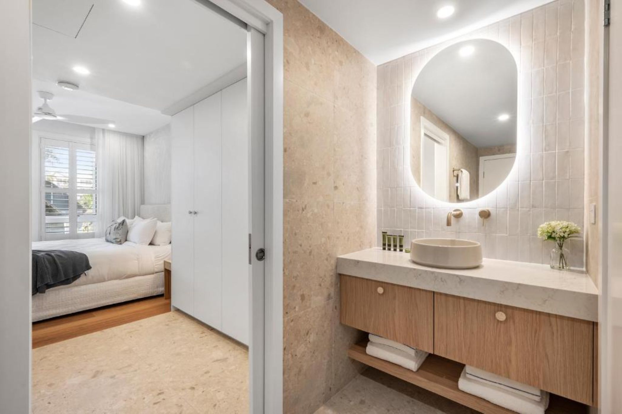 Une chambre avec un lit, une salle de bain avec un meuble-lavabo et un miroir, et un couloir.