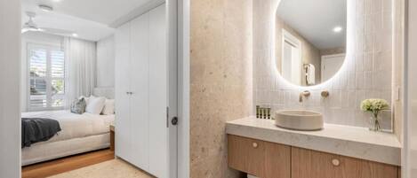 Une chambre avec un lit, une salle de bain avec un meuble-lavabo et un miroir, et un couloir.