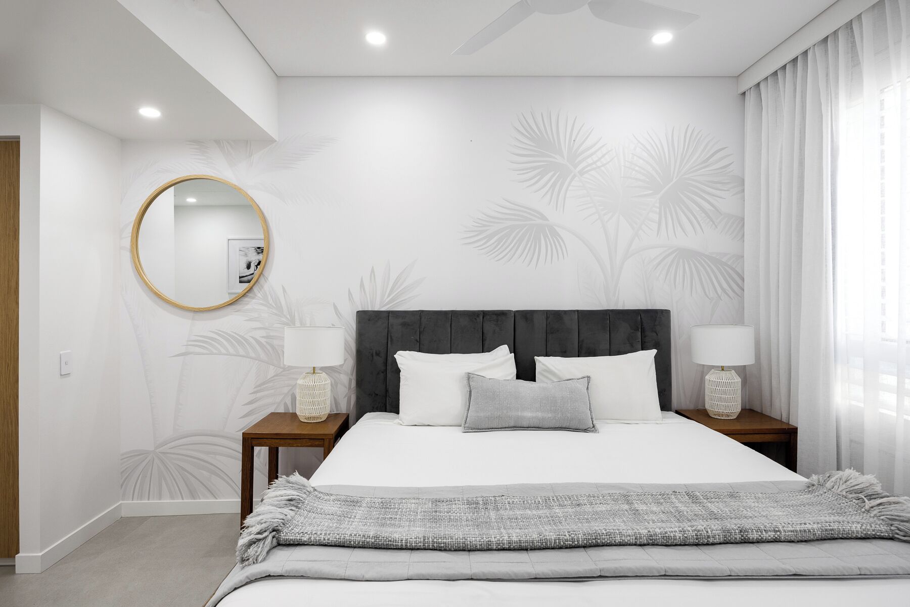 A modern bedroom with a large bed, two bedside tables, a round mirror, and a ceiling fan.