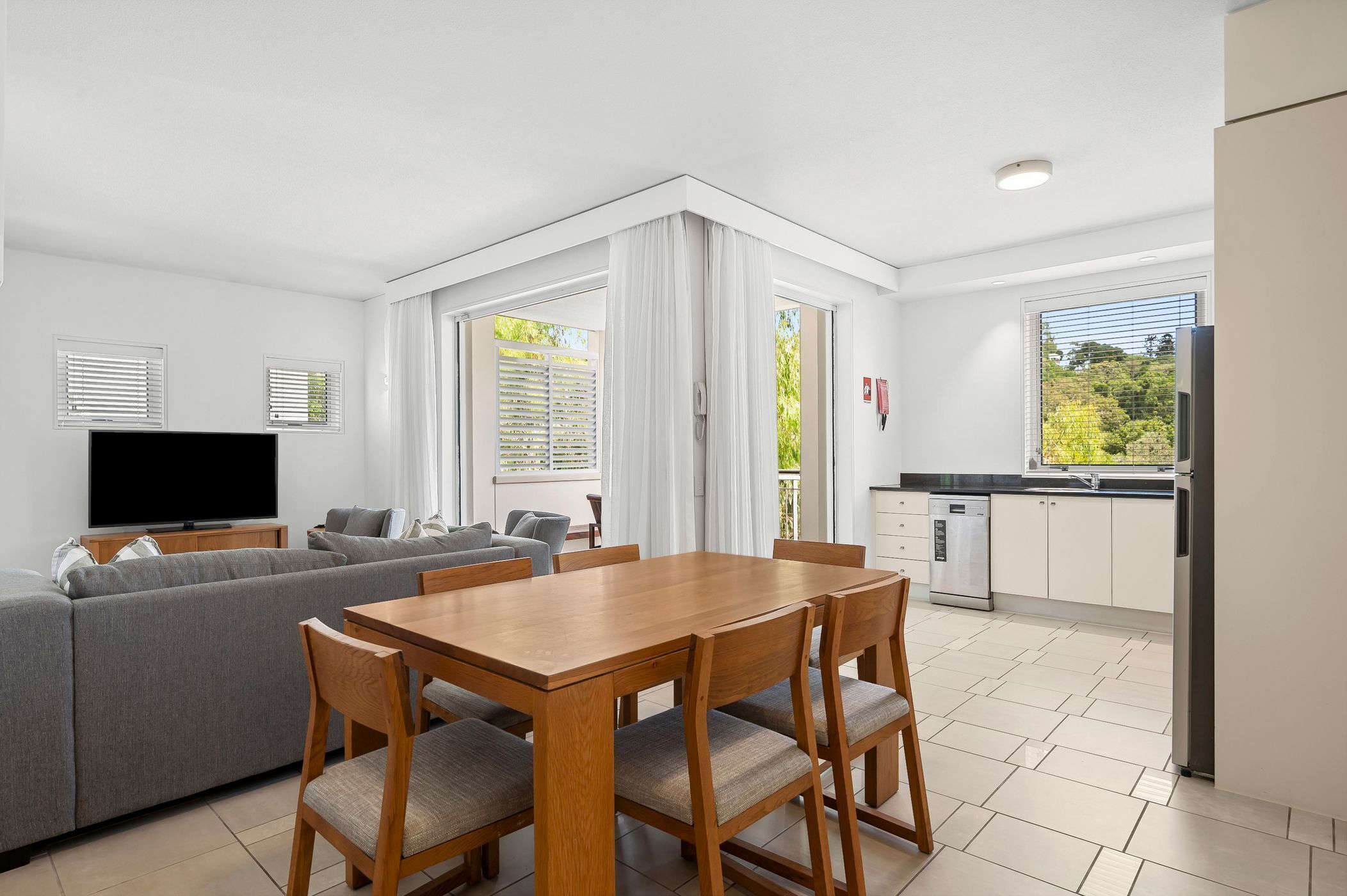 A modern living room with a grey sofa, wooden dining table, and chairs. There is a flat-screen TV on a stand, a kitchen area with a fridge, and a window with blinds.