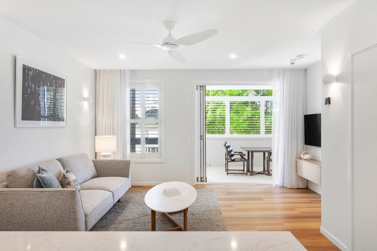 A modern living room with a sofa, coffee table, and a TV. There is a balcony with a view of greenery.