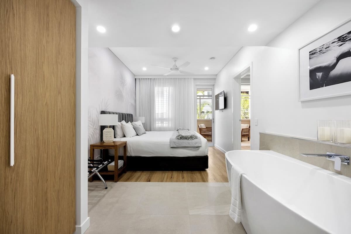 A modern bedroom with a large bed, a bedside table with a lamp, a ceiling fan, and a view of a balcony.