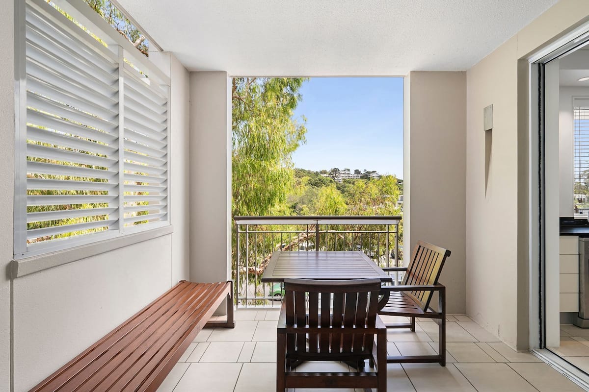 A balcony with a wooden table and chairs, a bench, and a view of greenery and buildings.