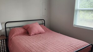 A bedroom with a bed, a nightstand, a wall-mounted air conditioner, a window with blinds, and a door.