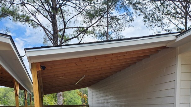 A covered outdoor area with a gravel floor, wooden beams, and a white wall.
