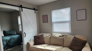 Ein Wohnzimmer mit einem beigen Sofa, zwei braunen Kissen, einer Schiebetür, einem Fenster mit Jalousien und zwei gerahmten Bildern an der Wand.