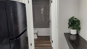 A narrow kitchen with a black refrigerator, a wooden floor, a white toilet, and a potted plant on a wooden shelf.