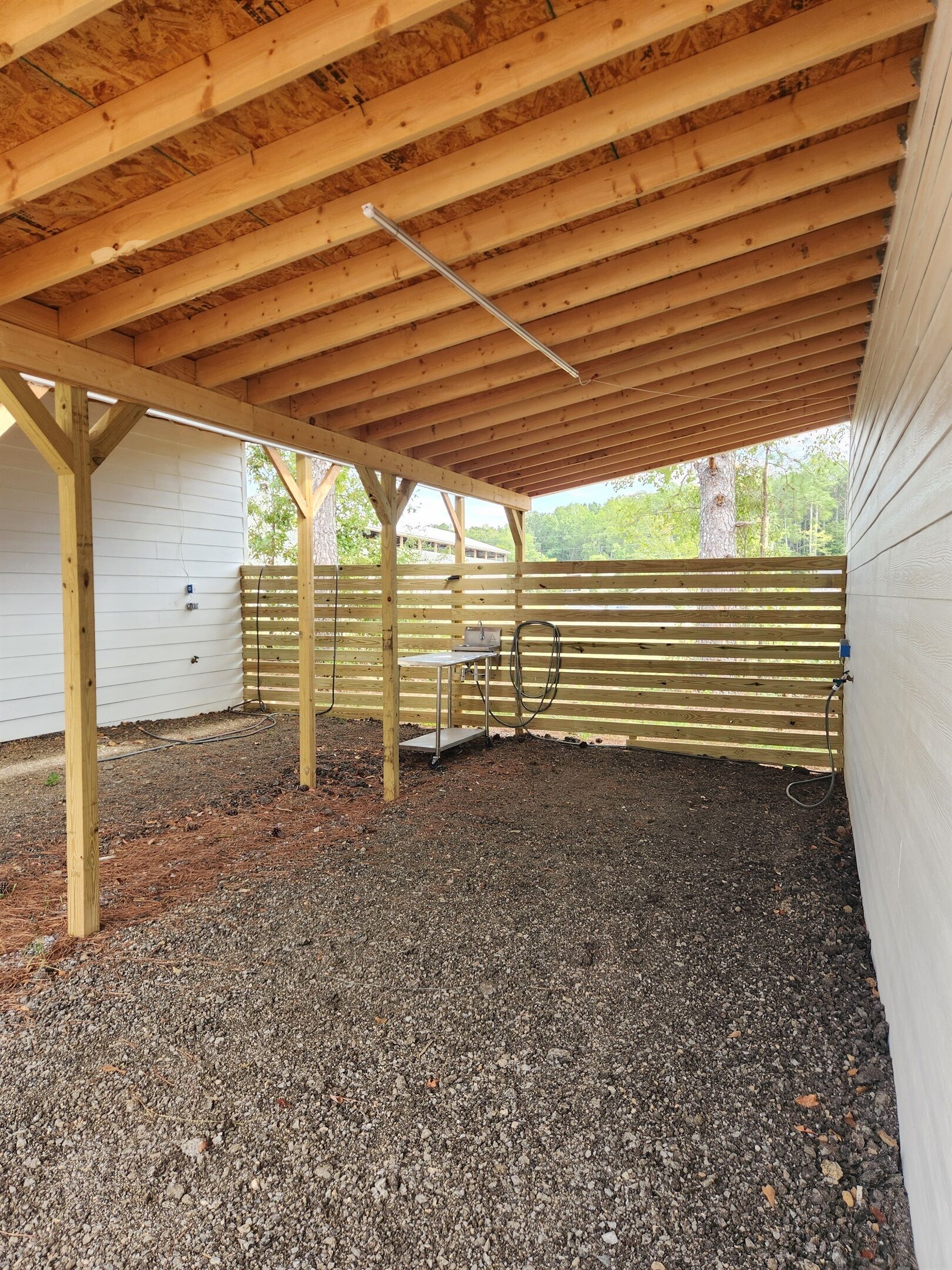An outdoor covered area with a gravel floor, wooden beams, and a metal table.