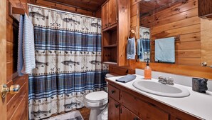 A wooden cabin bathroom with a plaid shower curtain, a white toilet, wooden cabinets, and a sink with a mirror above it. - Relax to Creek Sounds • Hot Tub Cabin • Firepit BBQ (Cherry Log)