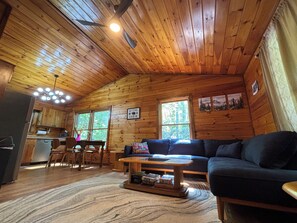 A cabin interior with a wooden ceiling, a sofa, a coffee table, a dining area, and a kitchen. - Relax to Creek Sounds • Hot Tub Cabin • Firepit BBQ (Cherry Log)