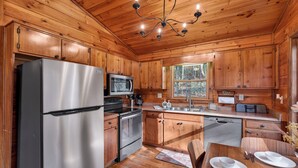 A modern kitchen with wooden cabinetry, stainless steel appliances, and a dining area with a wooden table and chairs. - Relax to Creek Sounds • Hot Tub Cabin • Firepit BBQ (Cherry Log)