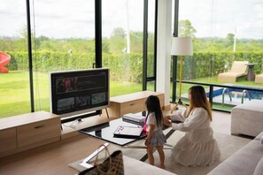 A modern living room with a large TV, a child playing, and a view of a pool and greenery. - Krajokkaew Mouthten Khao Yai (Pak Chong)