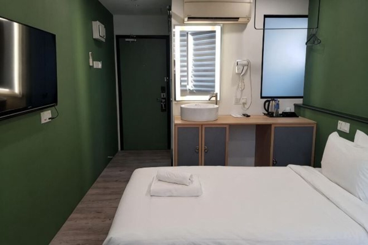 Een hotelkamer met een bed, nachtkastje met lamp, raam met zonwering, televisie aan de muur en een badkamer met wastafel en spiegel.