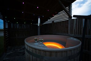 Un bain à remous en bois, avec deux verres de vin, entouré d’une terrasse en bois et d’une structure métallique agrémentée de lumières colorées.