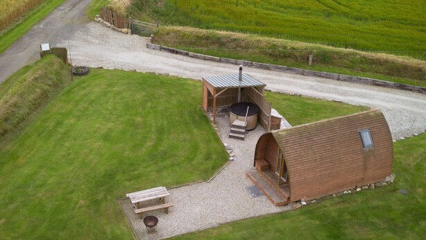 A rural property with a large open field, a gravel driveway, and a wooden structure with a hot tub.