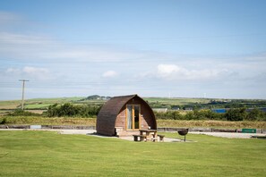 A small wooden cabin with a picnic table and a barbecue grill in a grassy field, surrounded by hills and a clear sky. - Ensuite Glamping Cabin With A View (Camelford)
