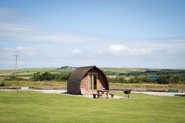 A small wooden cabin with a picnic table and a barbecue grill in a grassy field, surrounded by hills and a clear sky.
