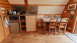 Eine kompakte Küche mit Mikrowelle, Wasserkocher und Spüle. Ein kleiner Kühlschrank und ein Esstisch mit Stühlen sind ebenfalls vorhanden. Der Raum hat Holzwände und eine schräge Decke.