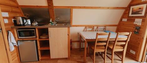 Cocina compacta con microondas, hervidor y lavabo. Hay un refrigerador pequeño y una mesa de comedor con sillas. La habitación tiene paredes de madera y techo inclinado.