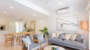 Una sala moderna con un sofá gris, una mesa de comedor de madera y sillas. Hay un cuadro grande en la pared, una lámpara de pie y una planta en maceta.