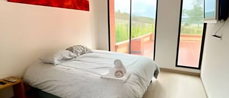 A modern bedroom with a wooden ceiling, a large bed, a nightstand, a TV, and a view of the outdoors.