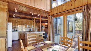 Interior de una cabaña de madera con una mesa de comedor, sillas, zona de cocina y un ventanal grande.