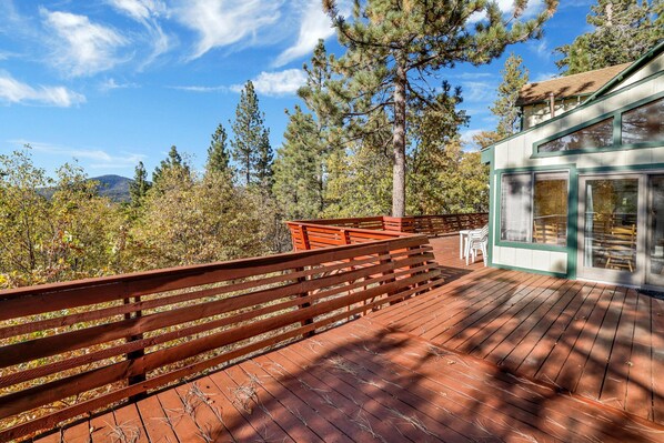 A wooden deck with a railing, overlooking a forested area with tall trees and a mountain in the distance.
