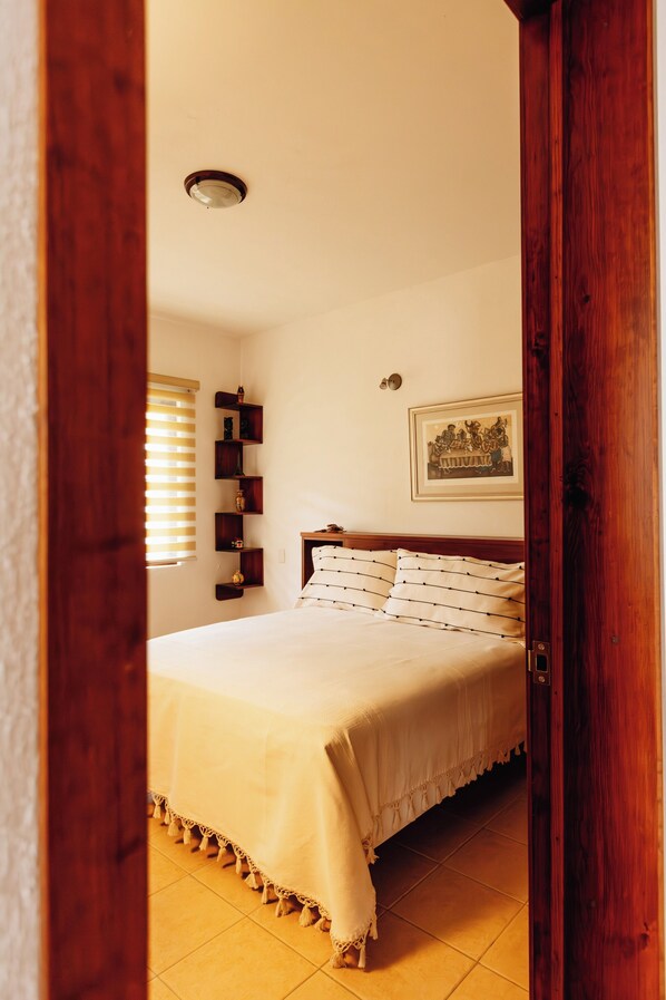 Un dormitorio con cama, cabecera de madera, repisa con objetos, un cuadro enmarcado y una ventana con persianas.