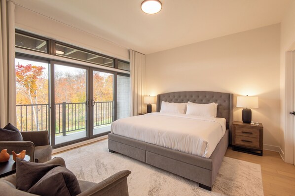A bedroom with a large bed, a nightstand, a chair, and a view of autumn trees through the sliding glass doors.