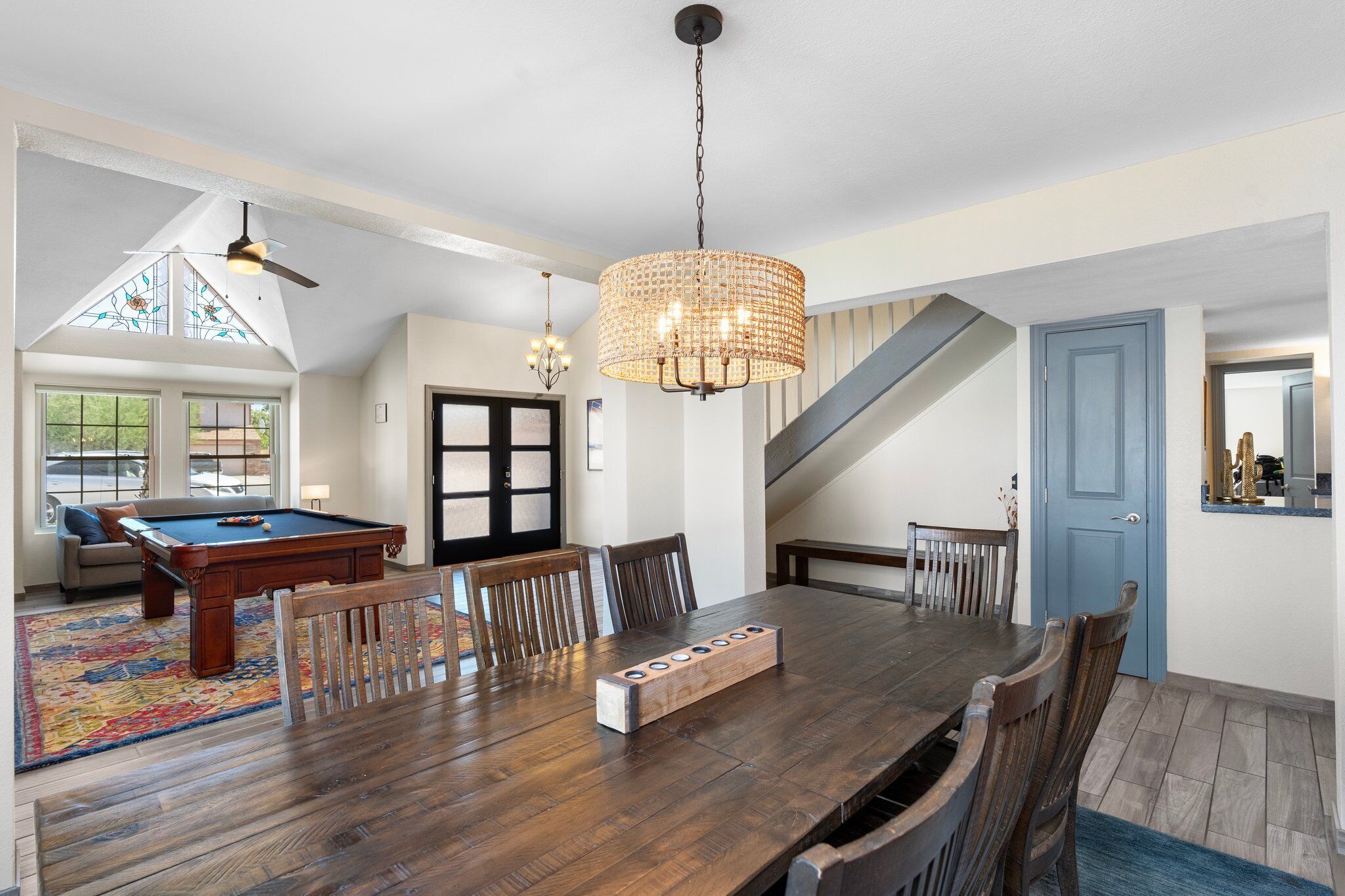 Ampla sala de jantar com mesa de madeira, cadeiras e um objeto decorativo central. Uma mesa de bilhar é visível ao fundo, e uma escadaria leva a outro andar.