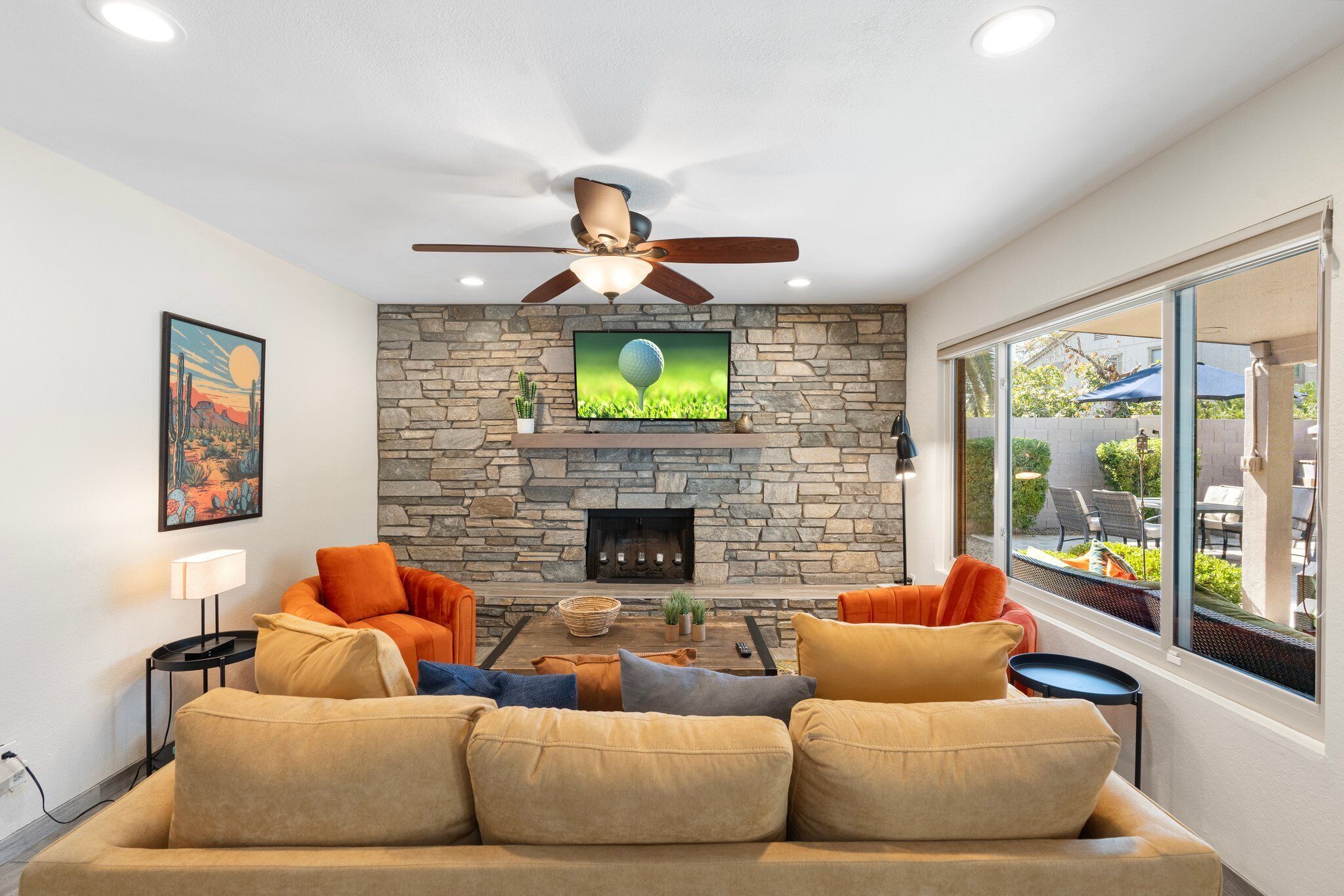 A living room with a stone fireplace, a ceiling fan, and a large window.