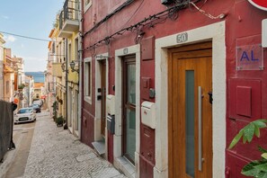 A narrow cobblestone street with red and yellow buildings, numbered doors, and a visible 'AL' sign.