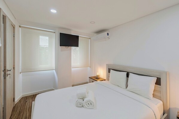 A hotel room with a large bed, a flat-screen TV mounted on the wall, a bedside table with a lamp, and a window with curtains.