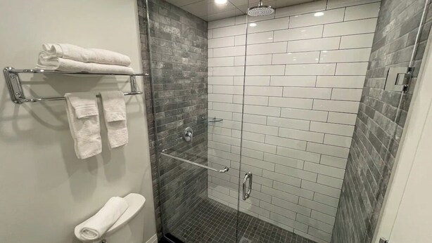 A modern bathroom with a glass-enclosed shower, white tiled walls, and a toilet with a towel rack. - Coastal Suites  (Grand Bend)