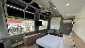 A modern bedroom with a large bed, a built-in bench, a small desk, and a window with a view of the outside. - Coastal Suites  (Grand Bend)