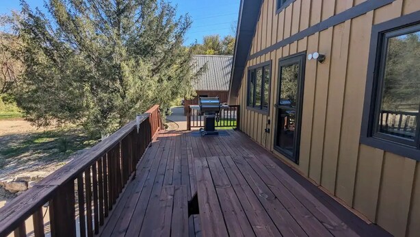 A wooden deck with a railing, a barbecue grill, and a view of a house with large windows. - A-frame home on Trout Stream (Stoddard)